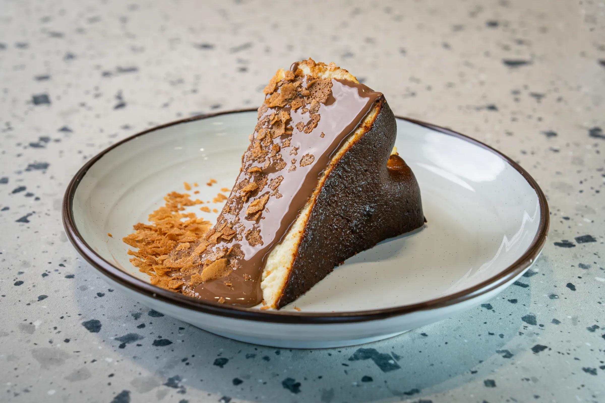 Slice tipped on its side, cut face under a thick chocolate glaze and biscuit crumb, burnt-dark crust behind, on a brown-rimmed plate